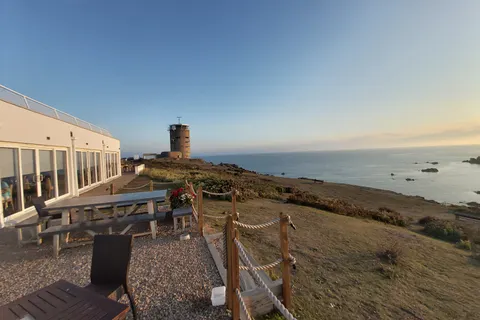 A pale building and wooden picnic tables overlook a coastal tower, grassy headland and sea at La Corbière.