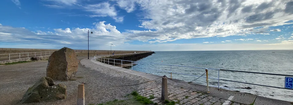 Stone paving and metal railings lead towards St Catherine's Breakwater with blue sea beneath streaked clouds.