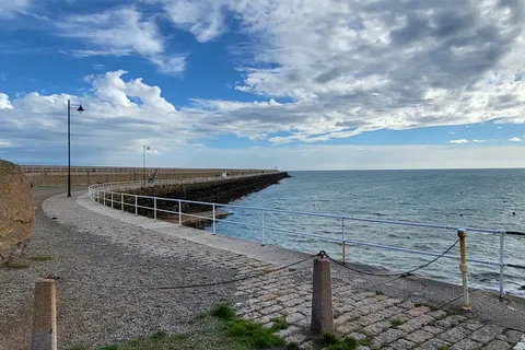 Stone paving and metal railings lead towards St Catherine's Breakwater with blue sea beneath streaked clouds.