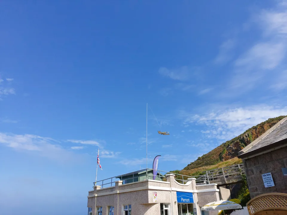 A seagull flies across a blue sky above small beach buildings and a green hillside at Plémont.