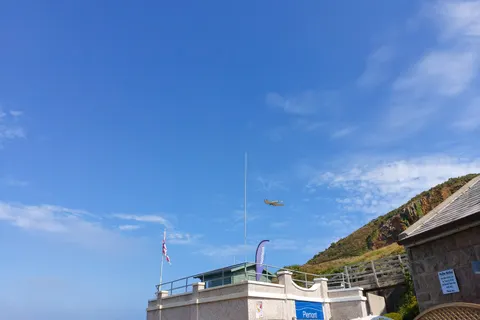 A seagull flies across a blue sky above small beach buildings and a green hillside at Plémont.