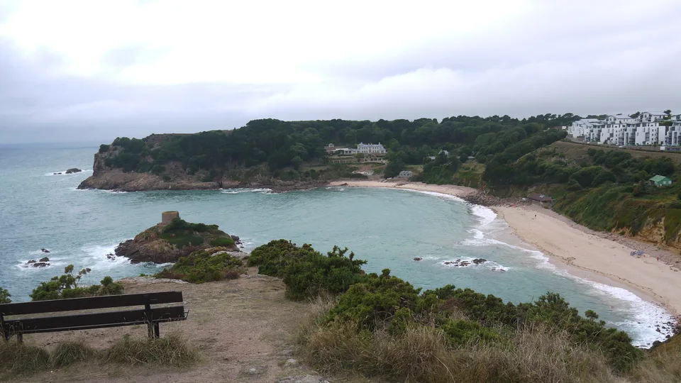 A sheltered sandy bay curves around a small rocky tidal islet, with wooded slopes and pale buildings above.