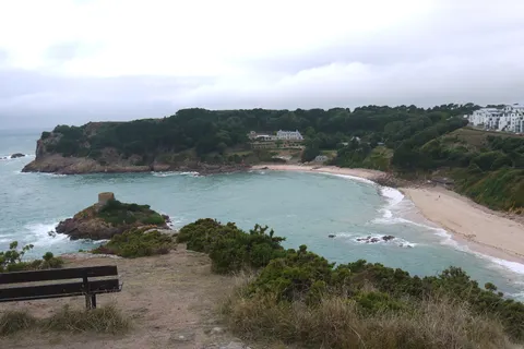 A sheltered sandy bay curves around a small rocky tidal islet, with wooded slopes and pale buildings above.