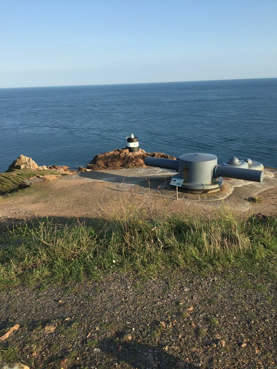 A small white coastal lookout and a low concrete structure stand on a grassy rocky headland above blue sea.
