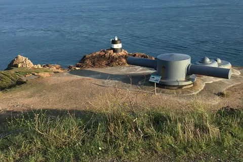 A small white coastal lookout and a low concrete structure stand on a grassy rocky headland above blue sea.