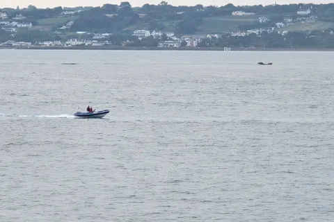 A small motorboat crosses grey-blue water with hillside homes on the distant shore of St Aubin's Bay.