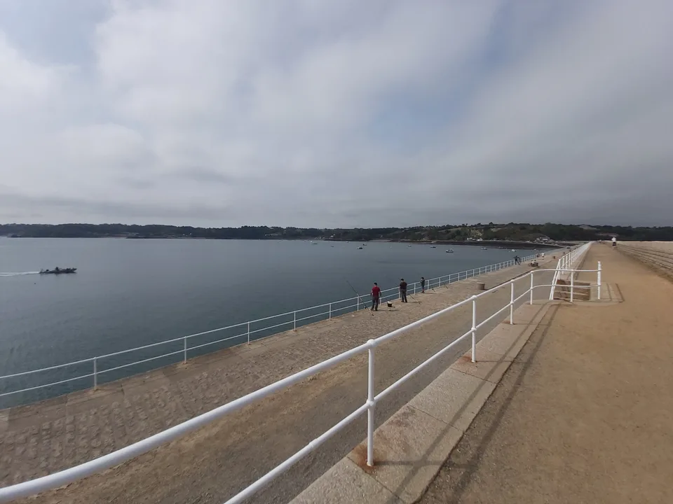 White railings line St Catherine's stone breakwater beside calm water and a distant wooded shore.