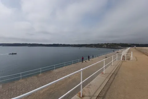White railings line St Catherine's stone breakwater beside calm water and a distant wooded shore.