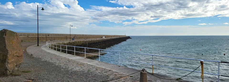 A wide coastal view shows St Catherine's stone breakwater, metal railings and blue sea beneath scattered clouds.