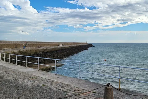 A wide coastal view shows St Catherine's stone breakwater, metal railings and blue sea beneath scattered clouds.