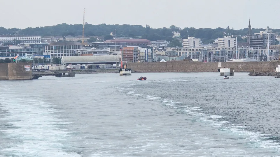 A foamy boat wake leads towards harbour structures and waterfront buildings at St Helier beneath a grey sky.