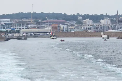 A foamy boat wake leads towards harbour structures and waterfront buildings at St Helier beneath a grey sky.