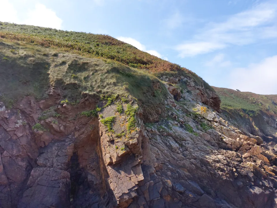 A steep pink-brown rock face rises below a grassy coastal slope with a glimpse of the sea at Plémont.