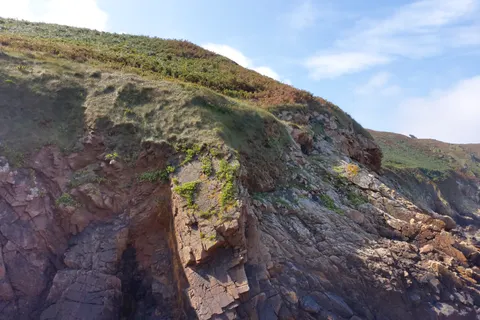 A steep pink-brown rock face rises below a grassy coastal slope with a glimpse of the sea at Plémont.