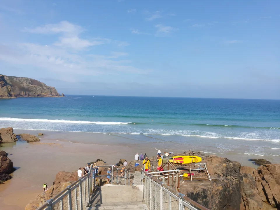 Metal handrails lead down steps towards Plémont beach, where small figures stand beside yellow kayaks.