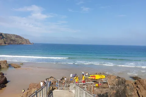 Metal handrails lead down steps towards Plémont beach, where small figures stand beside yellow kayaks.