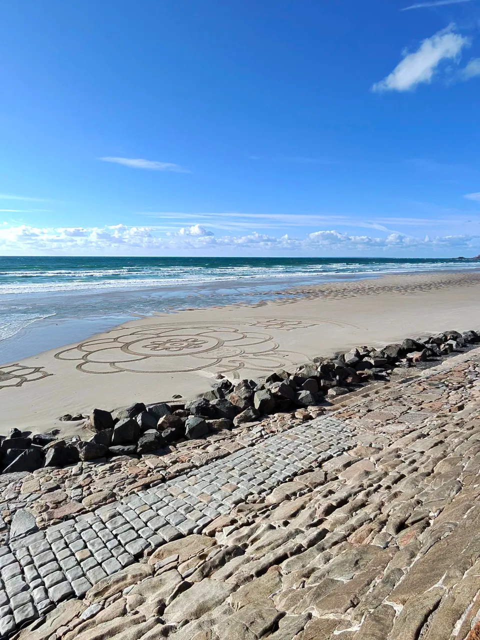 A sloping stone seawall borders pale sand and turquoise surf beneath blue sky at St Ouen's Bay.
