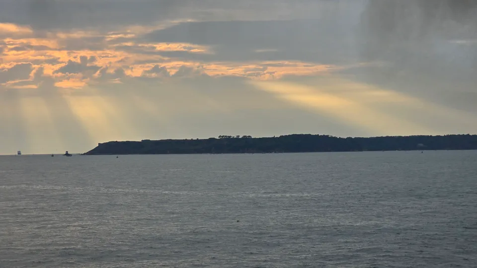Warm rays of sunlight break through grey clouds above the sea and a dark distant headland across St Aubin's Bay.