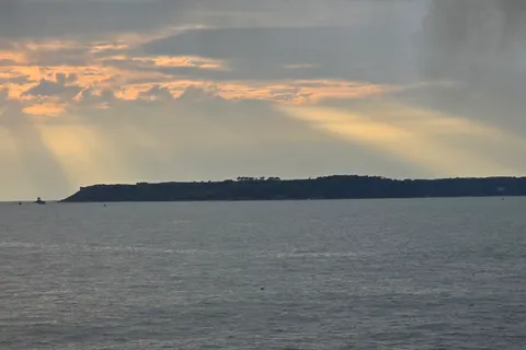 Warm rays of sunlight break through grey clouds above the sea and a dark distant headland across St Aubin's Bay.