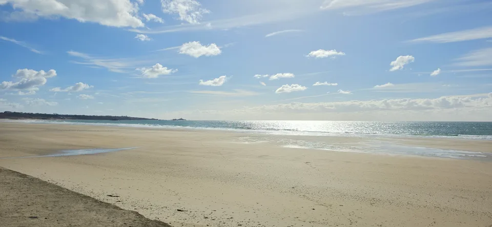 A wide pale sandy beach stretches beside sunlit sea at St Ouen's Bay beneath a lightly clouded blue sky.