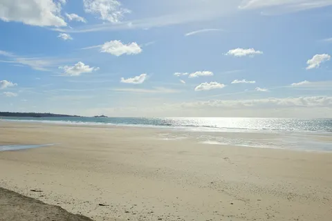 A wide pale sandy beach stretches beside sunlit sea at St Ouen's Bay beneath a lightly clouded blue sky.