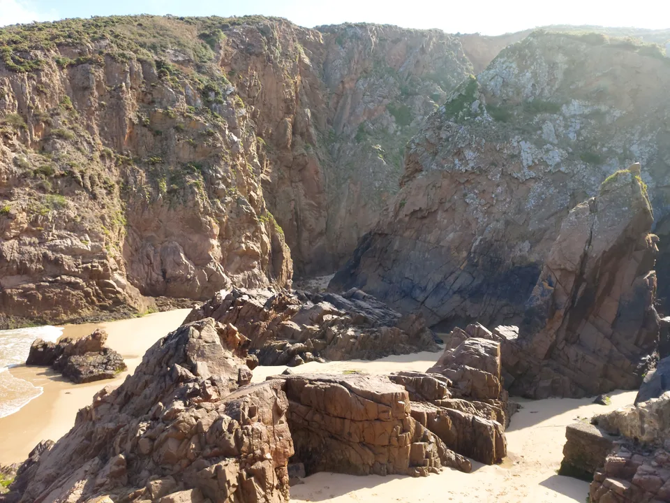 Sunlit boulders and a patch of sand sit beneath Plémont's steep cliffs beside a dark rocky recess.