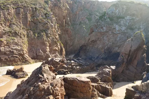 Sunlit boulders and a patch of sand sit beneath Plémont's steep cliffs beside a dark rocky recess.