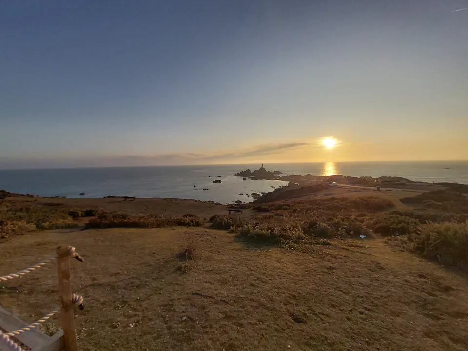 A grassy headland and coastal path overlook La Corbière Lighthouse and the low sun reflected across the sea.