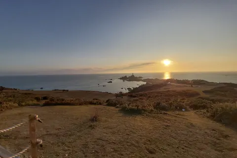 A grassy headland and coastal path overlook La Corbière Lighthouse and the low sun reflected across the sea.