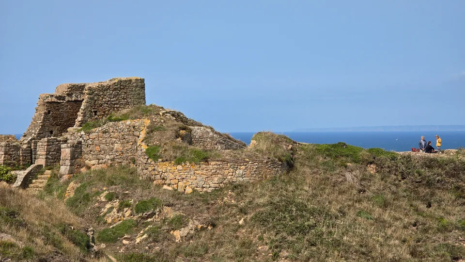 Sunlit stone castle walls stand on a rocky grassy headland at Grosnez with the blue sea behind.