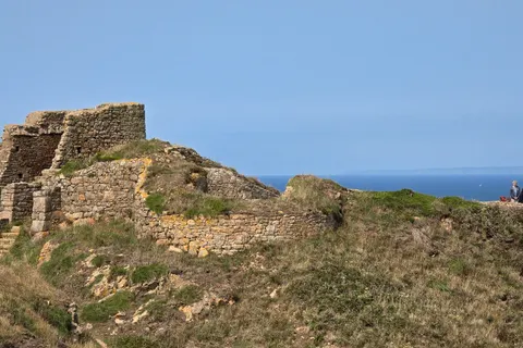 Sunlit stone castle walls stand on a rocky grassy headland at Grosnez with the blue sea behind.