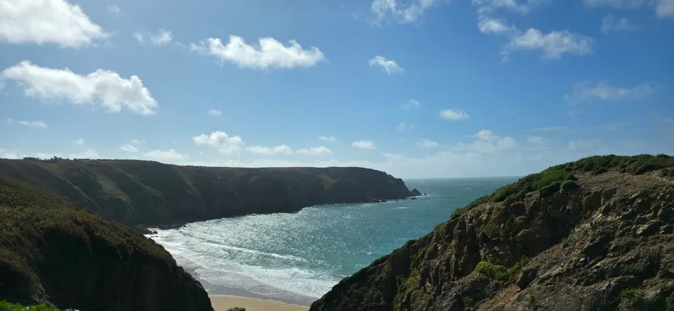 Steep green-topped cliffs frame sunlit blue water in a narrow coastal view near Plémont.