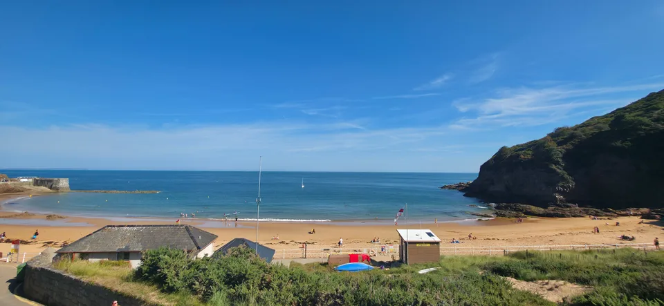 Beach buildings, pale sand and small boats line a sunny blue bay below a green headland at Grève de Lecq.