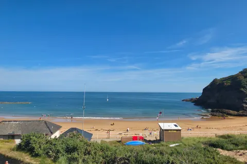 Beach buildings, pale sand and small boats line a sunny blue bay below a green headland at Grève de Lecq.