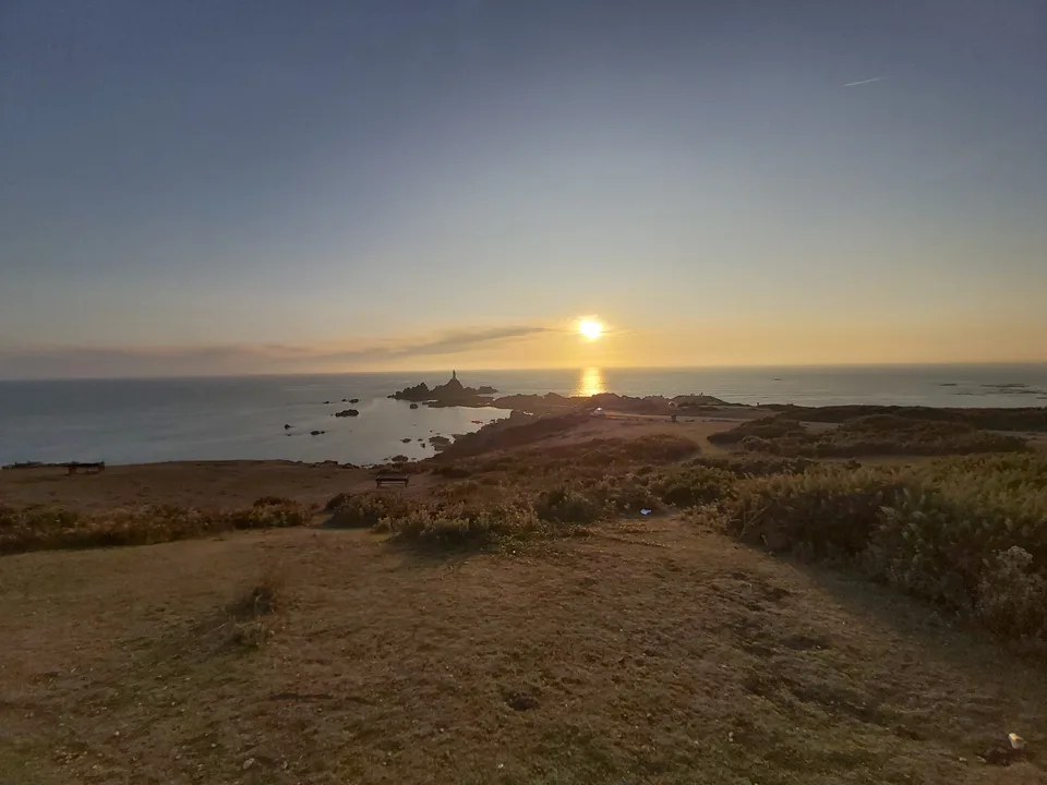 A low golden sun lights the sea beside La Corbière Lighthouse, with a path and rough grass in the foreground.
