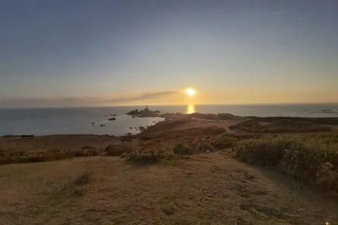A low golden sun lights the sea beside La Corbière Lighthouse, with a path and rough grass in the foreground.