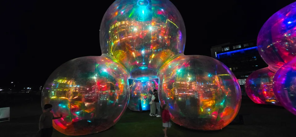 A group of transparent spheres glows with colourful reflections beside a building at night.