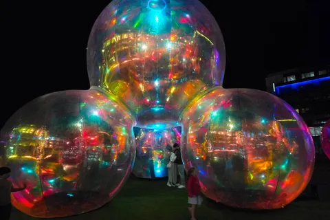 A group of transparent spheres glows with colourful reflections beside a building at night.