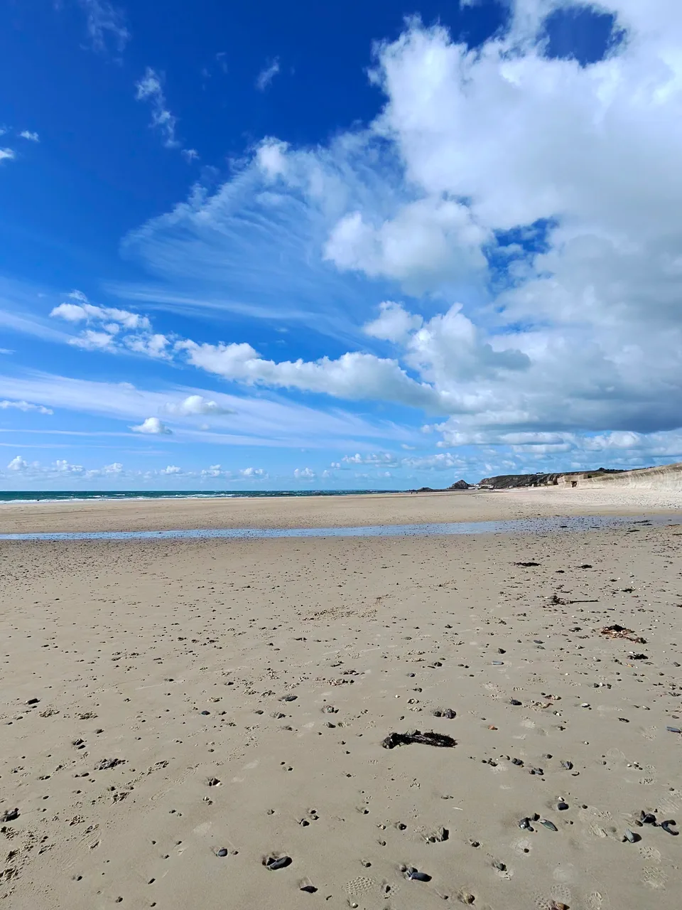 A vertical view shows pale sand, gentle surf and bright clouds above the sea at St Ouen's Bay.