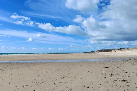 A vertical view shows pale sand, gentle surf and bright clouds above the sea at St Ouen's Bay.