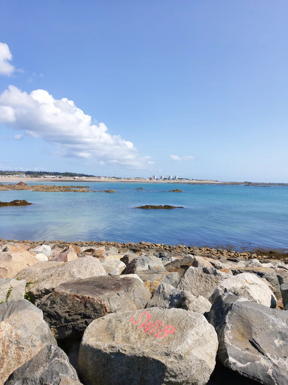 Large foreground rocks overlook blue water and a distant shoreline beneath white clouds across St Aubin's Bay.