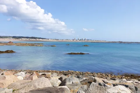 Large foreground rocks overlook blue water and a distant shoreline beneath white clouds across St Aubin's Bay.