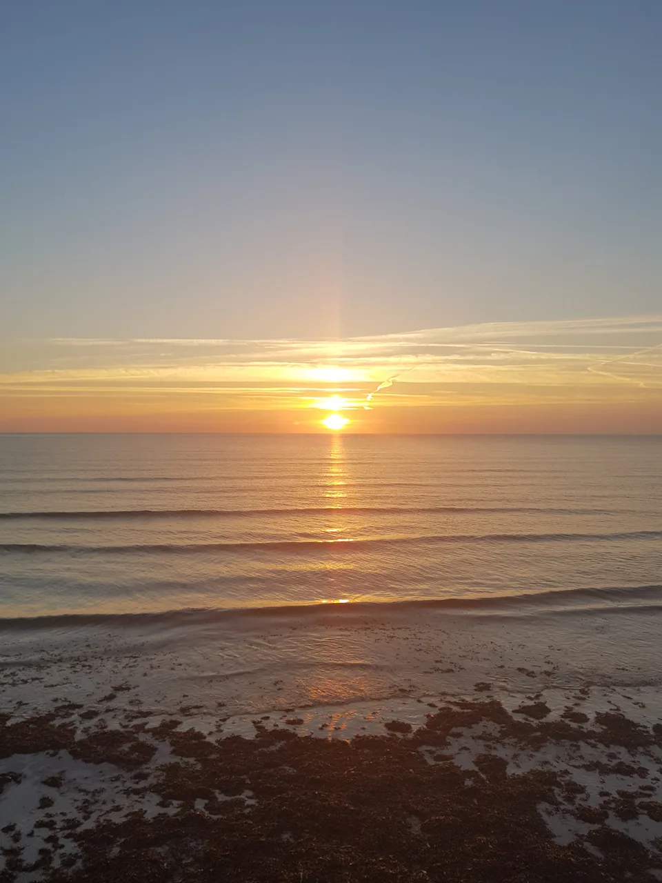 A golden sun and its reflection light the sea above a dark beach in a vertical coastal view.