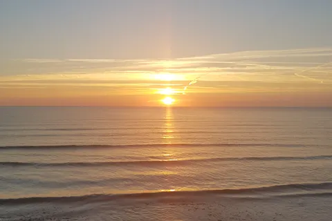 A golden sun and its reflection light the sea above a dark beach in a vertical coastal view.