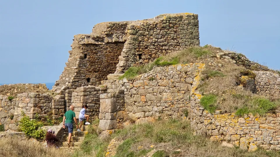 Two visitors stand near an opening in the sunlit stone ruins of Grosnez Castle on a grassy coastal slope.