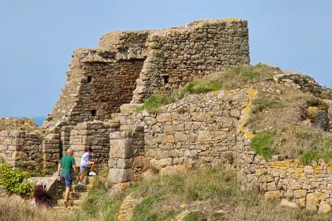 Two visitors stand near an opening in the sunlit stone ruins of Grosnez Castle on a grassy coastal slope.