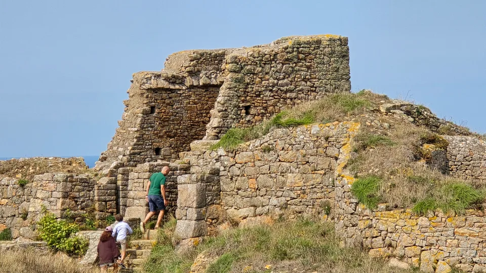 Visitors approach rough stone walls and a small opening among the ruins of Grosnez Castle under blue sky.