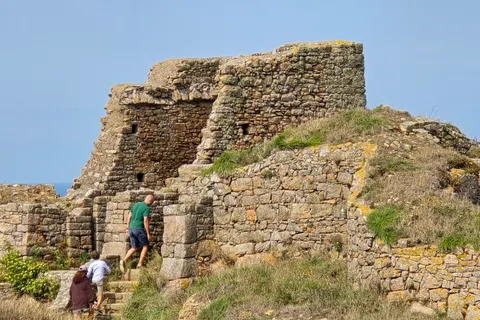 Visitors approach rough stone walls and a small opening among the ruins of Grosnez Castle under blue sky.