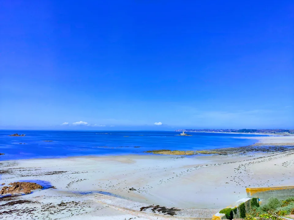 A strongly saturated blue sea and sky contrast with pale sand and dark shoreline rocks at St Ouen's Bay.