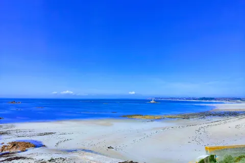 A strongly saturated blue sea and sky contrast with pale sand and dark shoreline rocks at St Ouen's Bay.